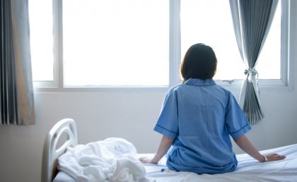 woman in blue shirt sits on a bed and looks out of a window